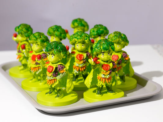 A group of small broccoli-themed figurines standing on round bases, arranged on a white tray against a light gray background.
