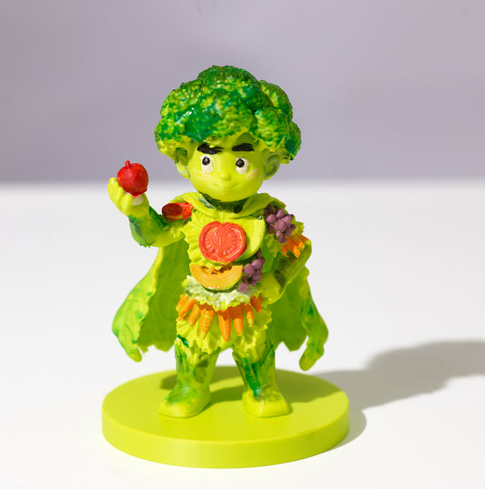 A small figurine of a broccoli character wearing a cape and holding a red apple, standing on a round yellow-green base against a plain light background.